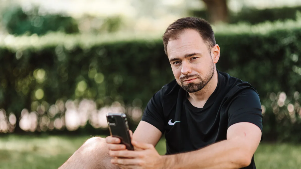 Mann sitzt draußen im Gras und blickt nachdenklich auf sein Smartphone